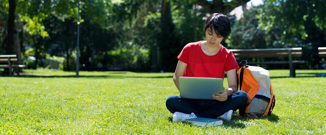 student wearing a red shirt sits with a laptop in the sunshine on a green lawn 