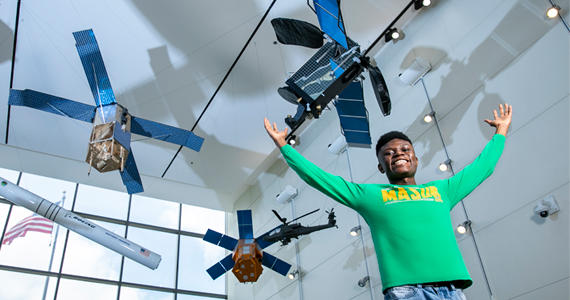 Sidney Boakye stands with his arms raised in front of several models of satellites. 