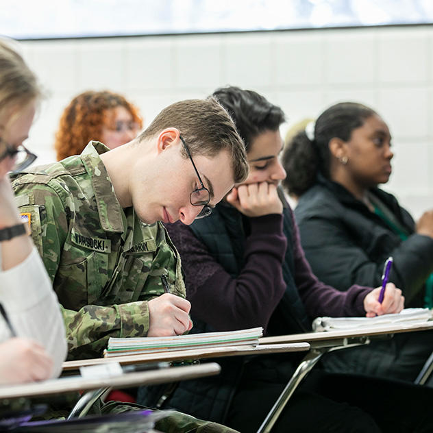 A Mason student wearing US Army camo fatigues sits in a classroom, head down, focused on what they're writing.
