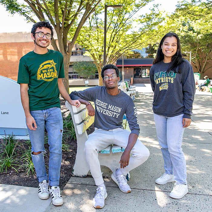 Three Mason students of different racial ethnicities are gathered in front of SUB I. They're wearing Mason sweatshirts and smiling at the camera.